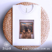 Футболка видатні українці, біла, на плоскій поверхні, з принтом, що зображує жінку, яка дивиться на групу чоловіків у картині, фон світло-сірий, деталі принта чіткі, зображення в золотій рамі, футболка розташована на плетеній основі.