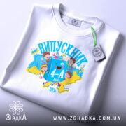 Футболка випускник молодшої школи, білого кольору, розкладена на поверхні. На фоні ніжно-фіолетовий колір. Принт яскравий, з малюнками дітей, книжок та написом 'ВИПУСКНИК 4-А клас', виконаний у синіх та жовтих тонах.