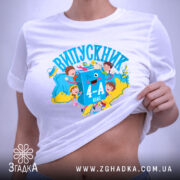 Футболка випускник молодшої школи, білого кольору, розкладена на поверхні. На фоні ніжно-фіолетовий колір. Принт яскравий, з малюнками дітей, книжок та написом 'ВИПУСКНИК 4-А клас', виконаний у синіх та жовтих тонах.