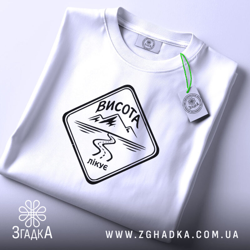 Футболка висота лікує. Складена біла футболка з чорним принтом, на якому напис 'ВИСОТА лікує', фон світло-сірий, видно етикетку з зеленим шнурком.