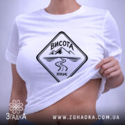 Футболка висота лікує. Складена біла футболка з чорним принтом, на якому напис 'ВИСОТА лікує', фон світло-сірий, видно етикетку з зеленим шнурком.