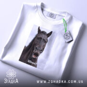 Футболка з мемним конем, білого кольору, складена на поверхні. Принт з конем розташований по центру, на ньому написано 'Шо?'. На футболці є етикетка з зеленим шнурком. Фон світло-сірий, що створює контраст з футболкою.