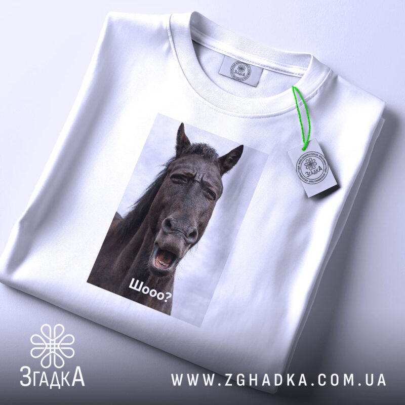 Футболка з мемним конем, білого кольору, складена на поверхні. Принт з конем розташований по центру, на ньому написано 'Шо?'. На футболці є етикетка з зеленим шнурком. Фон світло-сірий, що створює контраст з футболкою.