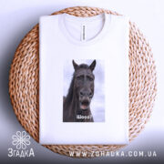 Футболка з мемним конем, білого кольору, складена на плетеній кошику. Принт з конем розташований по центру, на ньому написано 'Шо?'. Фон світло-сірий, а кошик додає текстурності до зображення.