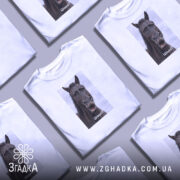 Футболка з мемним конем, білого кольору, зображена на молодій людині, яка дивиться в камеру. Принт з конем займає центральну частину, на ньому написано 'Шо?' у нижній частині. Фон світло-фіолетовий, що підкреслює яскравість футболки.