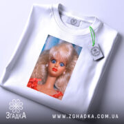 Футболка з мемним обличчям барбі, біла, на складеній футболці, фон світло-фіолетовий, принт зображує барбі з великими блакитними очима та світлим волоссям, одягнена в червону сукню, на футболці є етикетка.