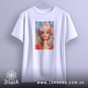 Футболка з мемним обличчям барбі, біла, підвішена на вішалці, фон світло-сірий, принт зображує барбі з великими блакитними очима та світлим волоссям, одягнена в червону сукню.