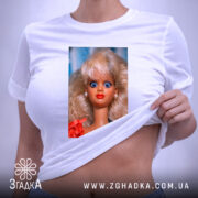Футболка з мемним обличчям барбі, біла, на складеній футболці, фон світло-фіолетовий, принт зображує барбі з великими блакитними очима та світлим волоссям, одягнена в червону сукню, на футболці є етикетка.