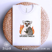 Футболка зазбогійся з лемуром, біла, складена на плетеній основі, фон світло-сірий, принт з лемуром з червоними очима та вогнем.