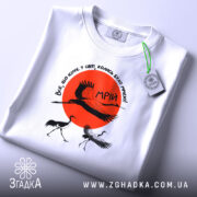 Футболка журавлі та мрія, біла футболка, зображена на столі, ракурс зверху, фон світло-сірий, принт з червоним сонцем та журавлями, на футболці є етикетка.