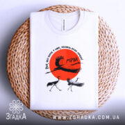 Футболка журавлі та мрія, біла футболка, зображена на плетеному кошику, ракурс зверху, фон світло-сірий, принт містить червоне сонце та журавлів, текст на футболці українською мовою.