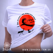 Футболка журавлі та мрія, біла футболка, зображена на столі, ракурс зверху, фон світло-сірий, принт з червоним сонцем та журавлями, на футболці є етикетка.