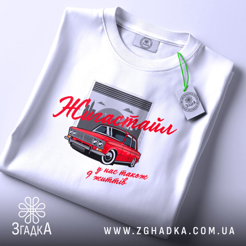 Футболка жигастайл білого кольору, зображена на плоскій поверхні. Принт з червоним автомобілем та написом 'Жигастайл' у червоному кольорі, розташований на фоні сірого гаража. На футболці видно етикетку з зеленим шнурком.