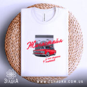 Футболка жигастайл білого кольору, складена на плоскій поверхні з плетеного матеріалу. Принт з червоним автомобілем та написом 'Жигастайл' у червоному кольорі, фон зображує сірі хмари та гаражні ворота.