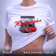 Футболка жигастайл білого кольору, зображена на плоскій поверхні. Принт з червоним автомобілем та написом 'Жигастайл' у червоному кольорі, розташований на фоні сірого гаража. На футболці видно етикетку з зеленим шнурком.