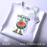 Футболка Жирафа Селесте на білому фоні, зображена в плоскому вигляді, принт з жирафом у космічному шоломі на кавуні, з яскравими оранжевими черевиками, на етикетці видно логотип бренду, фон світло-сірий.