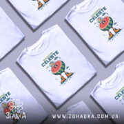 Футболка Жирафа Селесте на білому фоні, зображена на чоловікові, який позує в три чверті, принт з жирафом у космічному шоломі, що стоїть на кавуні, з яскравими оранжевими черевиками, фон світло-сірий.