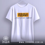 Футболка зроблю за годину. Біла футболка, підвішена на вішалці, з коротким рукавом. Принт на грудях чорного та оранжевого кольорів, текст 'ЗРОБЛЮ ЗА ГОДИНУ ...ПРОТЯГОМ ТИЖНЯ'. Фон світло-сірий, з м'яким освітленням.