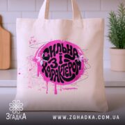 Шопер сильна і з характером, бежевий, на кухонному фоні, яскравий принт, бренд.