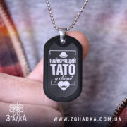 Жетон найкращий тато, чорного кольору, матова поверхня, зображений в руці, фон з розмитими кольорами, видно частину одягу на фоні.