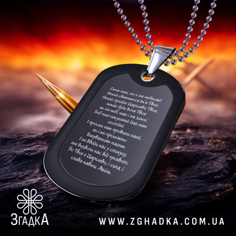 Жетон Отче наш, чорного кольору, матова поверхня, зображений з бокового ракурсу на фоні з червоними та чорними відтінками, видно текст молитви на жетоні та металевий ланцюжок.