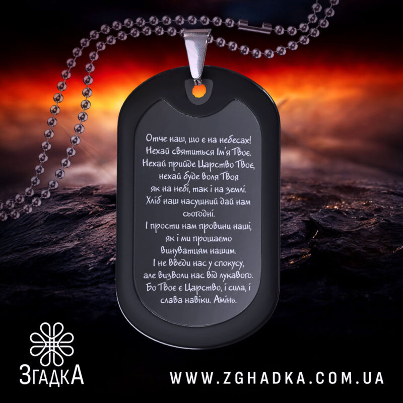 Жетон Отче наш, чорного кольору, матова поверхня, зображений з верхнього ракурсу на фоні з червоними та чорними відтінками, видно текст молитви на жетоні та металевий ланцюжок.