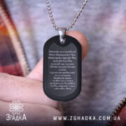 Жетон Отче наш, чорного кольору, матова поверхня, зображений з близького ракурсу в руці, на фоні з розмитими кольоровими відтінками, видно текст молитви на жетоні та металевий ланцюжок.