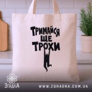 Шопер тримайся ще трохи, бежевий, на столі, з рослиною на фоні, бренд.