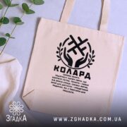 Шопер Колард слов’янський оберіг бежевий, з чорним принтом, на столі з рослинами,.